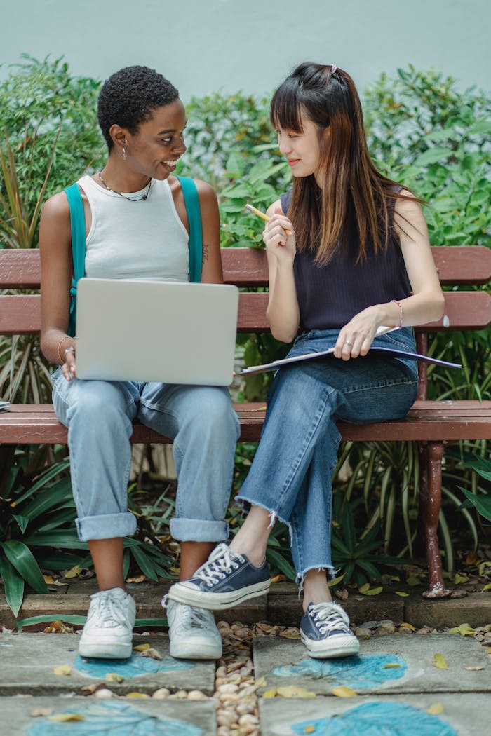 Two young women studying with a laptop and notebook on a park bench.